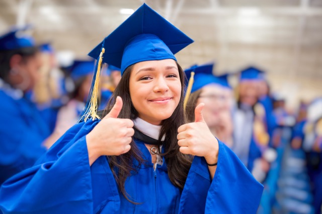 Young college girl in cap and gown at the graduation ceremony giving the thumbs up for success.