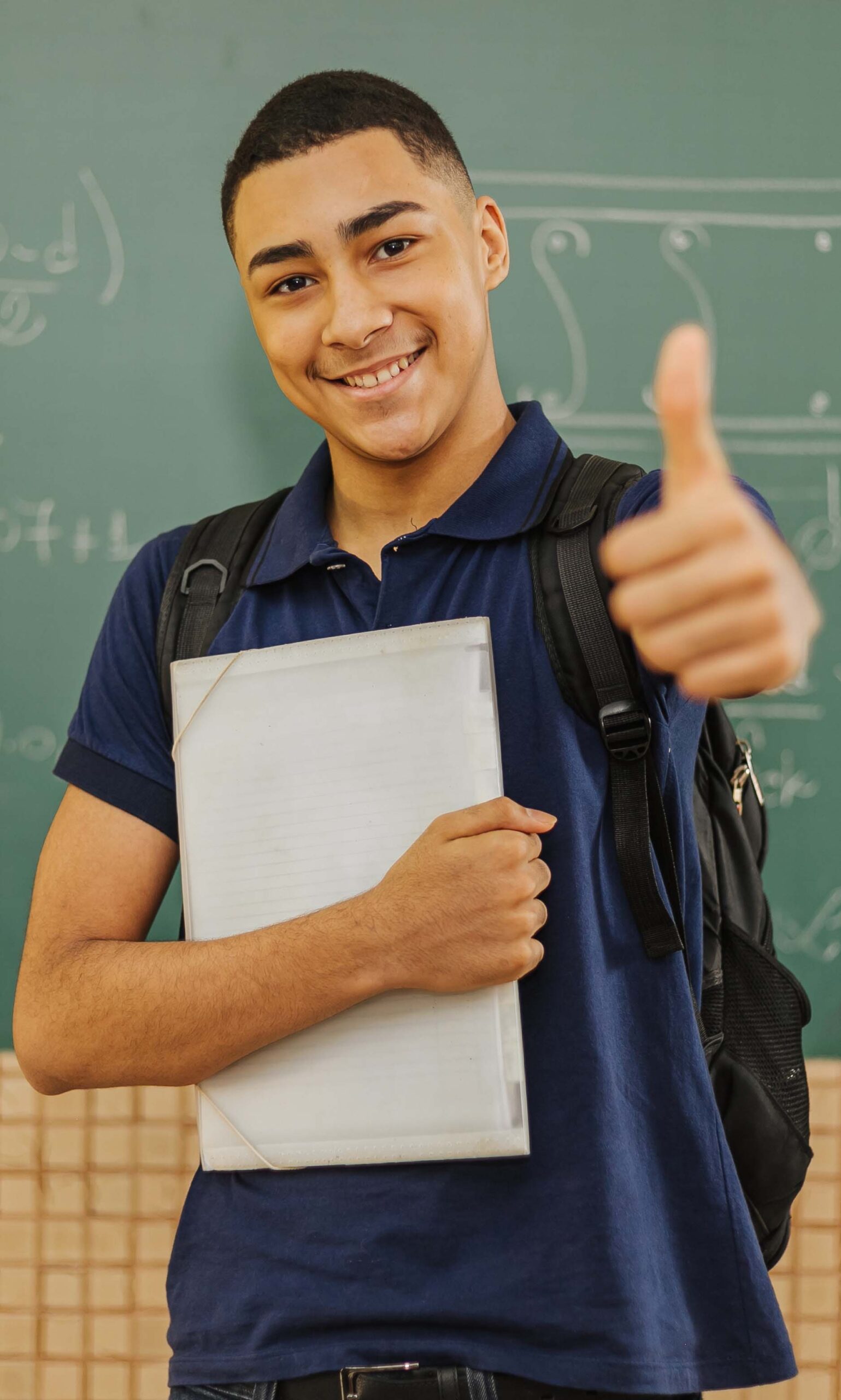 Latin men student smiling wearing backpack holding a notebook in a classroom with Thumbs up